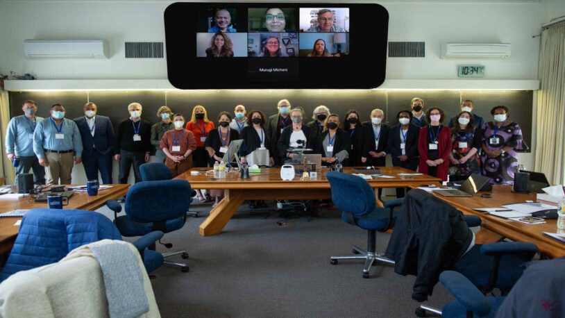 A photograph of 22 people standing in two rows in front of the Banbury Center. All meeting participants are wearing masks and blue lanyards with name tags. There are seven people displayed on the projector, smiling.