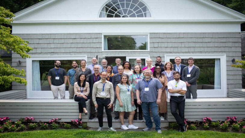 A photograph of 23 people standing in three rows on the wood back patio of the Banbury Center's conference room. All meeting participants are wearing blue lanyards with name tags and are smiling.